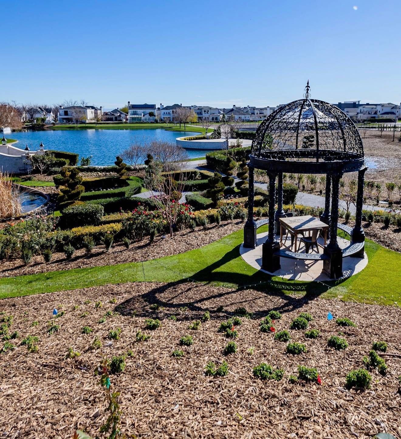 A wrought iron gazebo at the centre of a clipped rose parterre beside the lake
